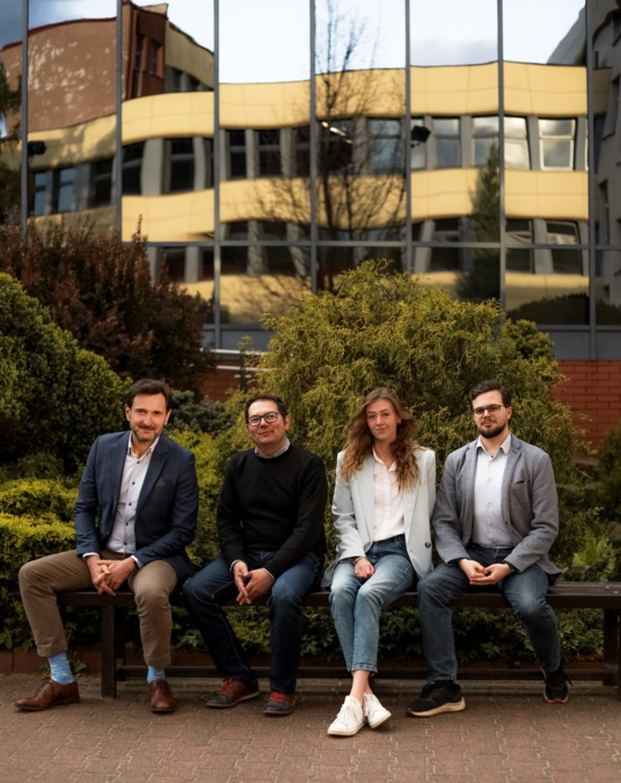 Cztery osoby siedzące na ławce na tle zieleni i budynku